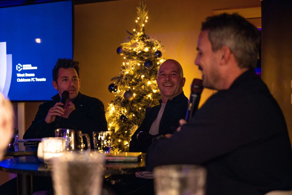 De drie organisatoren zitten pratend aan een tafel met een kerstboom achter hen bij het evenement van De Voetbalbondgenoten.