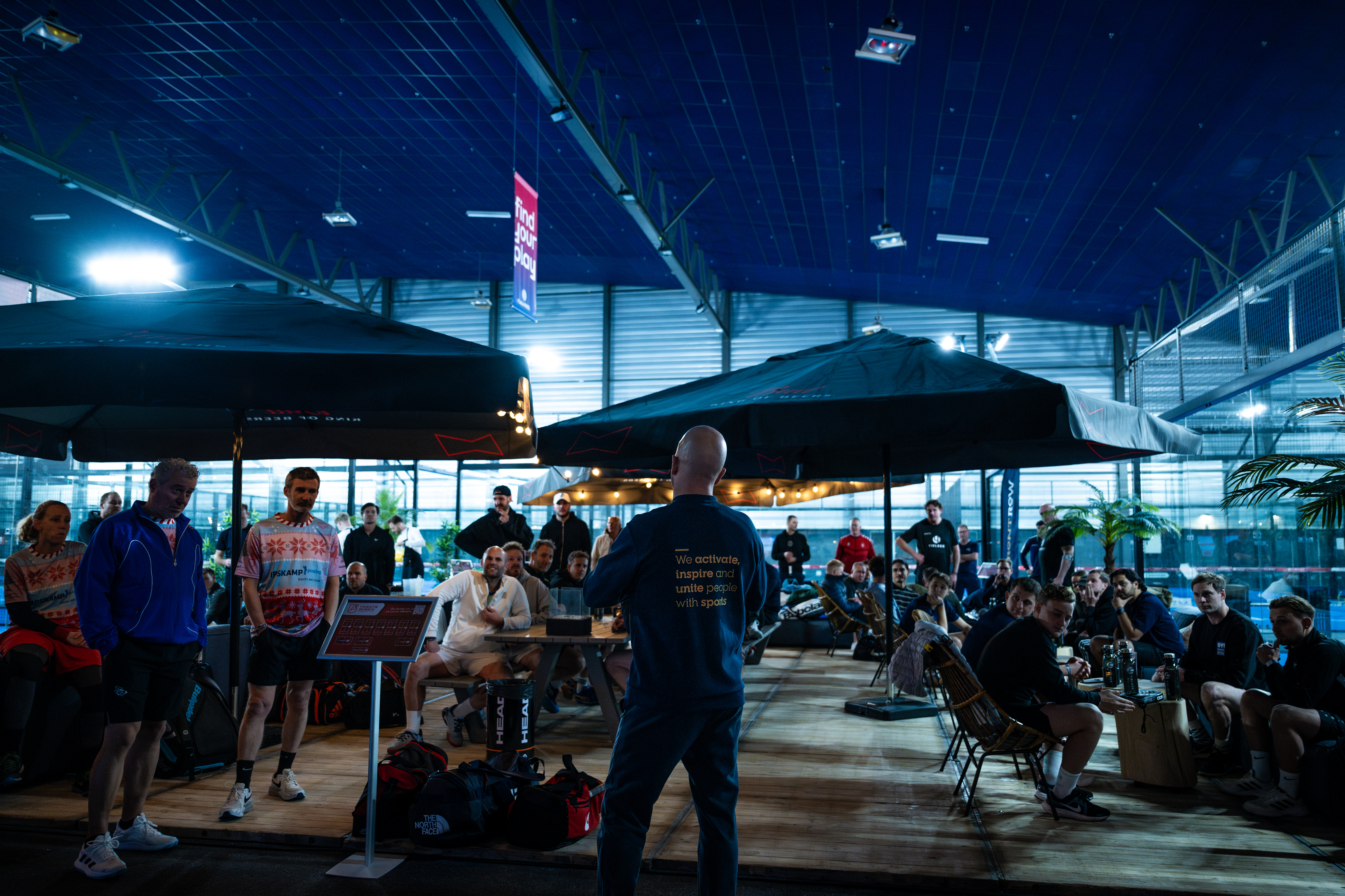 Deelnemers aan het indoor padel event Twents Ross'n zitten aan tafels onder parasols en luisteren naar de organisator van het event.