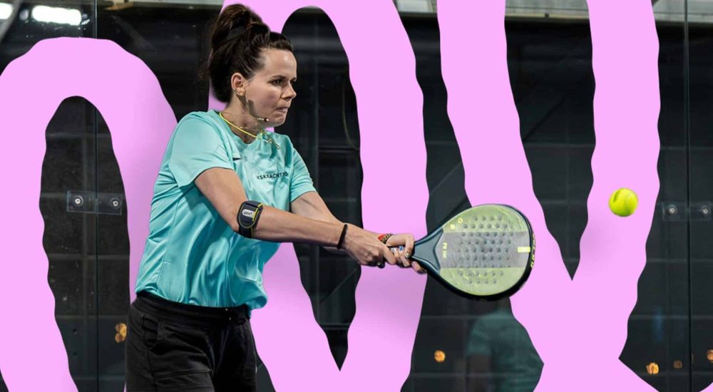Vrouw in een lichtblauw sportshirt slaat een padelbal tijdens een wedstrijd op een baan met roze achtergrond.